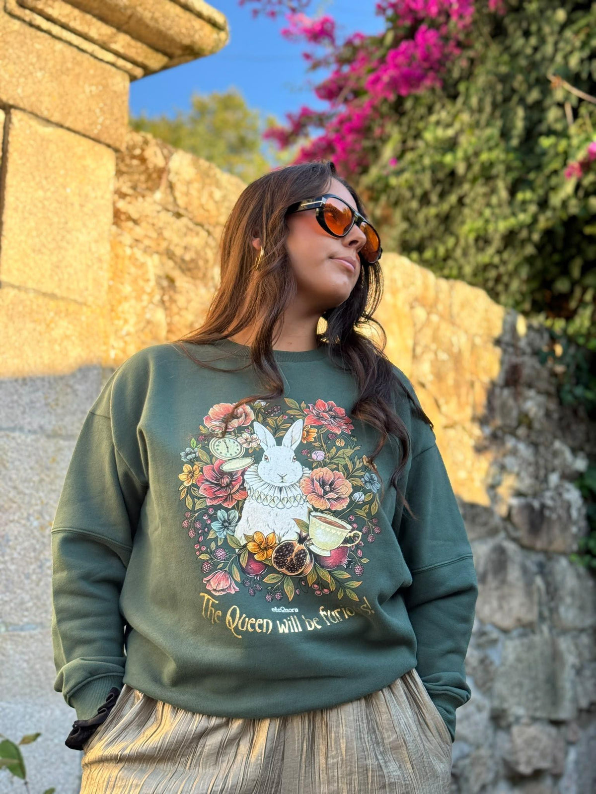 Mujer con sudadera verde eleOnora con estampado ilustrado de un conejo blanco entre flores y el texto 'The Queen will be furious!'